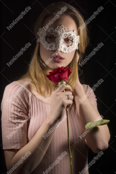 Portrait of a shy girl wearing lace mask and red rose on black background