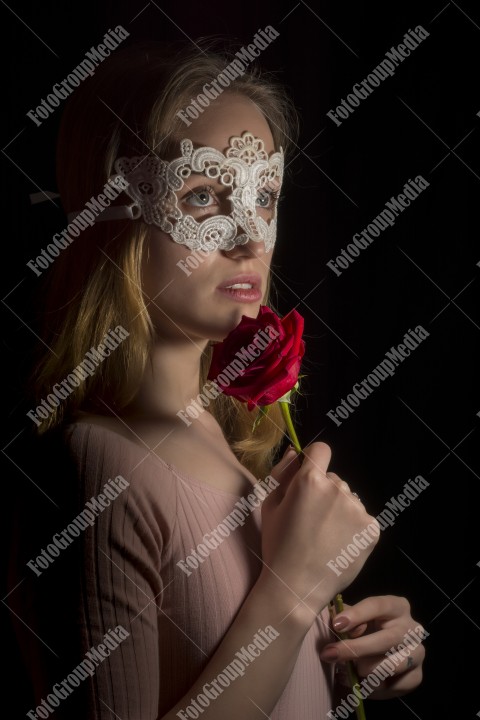 Portrait of a shy girl wearing lace mask and red rose on black background