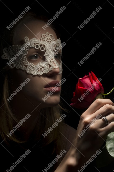 Portrait of a shy girl wearing lace mask and red rose on black background