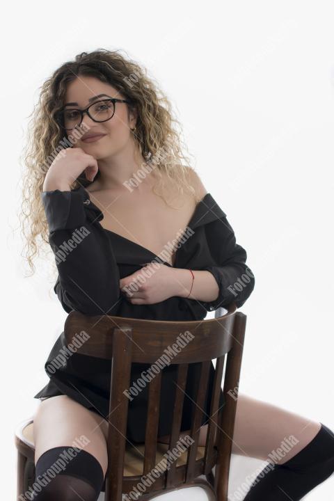 Curly blond long hair in black jacket and stocking posed on chair at studio against white background.